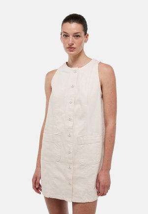 Femme aux cheveux foncés portant une robe beige clair sans manches, boutonnée devant avec deux poches devant, debout sur un fond clair uni.