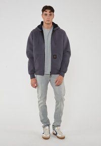 Felpa grigia con zip e interno nero, abbinata a una maglietta grigia, jeans chiari e sneakers bianche con dettagli blu navy e gialli.