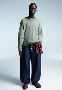 Helles graues Rippenoberteil, lockere dunkelblaue Jeans mit Knopfdetail und ein rotes Halstuch, das um die Taille gebunden ist; dunkelburgunderfarbene Schuhe.