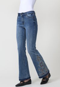 Jeans de mezclilla azul de tiro alto y acampanados con bordado floral colorido en el dobladillo y un borde deshilachado. Cuenta con bolsillos frontales y un cierre de botones clásico.