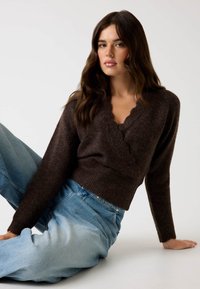 Maglione avvolgente lungo marrone, con scollo ondulato e texture, abbinato a jeans alta vita azzurri. Tessuto morbido, vestibilità comoda.