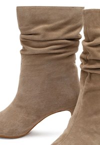 Stivaletti alla caviglia in suede beige con un design morbido, dotati di un tacco affusolato e una texture liscia. Nessuna hardware o pattern visibili.