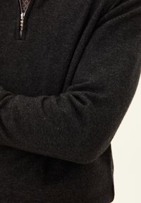 Dunkelgrauer Strickpulloverärmel mit einem silbernen Reißverschlussdetail Nähe des Kragens, das subtile Textur und Falten im Stoff zeigt.