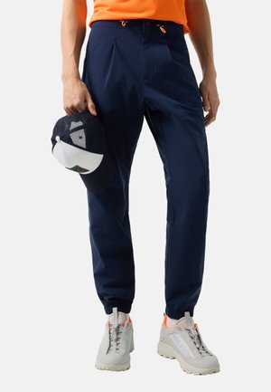 Persoon draagt marineblauwe joggingbroek, witte en oranje sneakers, houdt een marineblauw-witte baseballpet naast zich.