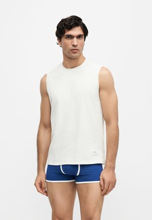 Hombre con cabello corto y oscuro que lleva una camiseta blanca sin mangas y calzoncillos bóxer azules con ribete blanco, de pie contra un fondo liso.