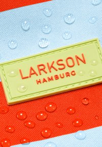 Primo piano di un tessuto impermeabile con gocce d'acqua e un'etichetta verde con la scritta "LARKSON HAMBURG" cucita su materiale rosso e azzurro chiaro.