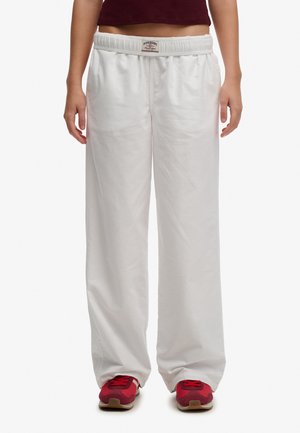 Persona che indossa pantaloni bianchi larghi con vita elastica e sneakers rosse, in piedi davanti a uno sfondo uniforme.