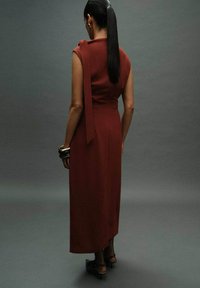 Langes, ärmelloses Kleid aus tiefrotem Stoff, mit einem Rückenschnürdetail und einer taillierten Silhouette. Getragen mit klobigen silbernen Armreifen und High-Heels.