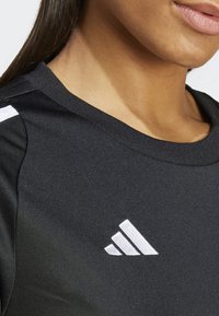 Tricou sport negru cu guler rotund, textură netedă, și trei dungi albe pe umărul stâng, având un logo brodat.