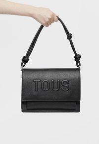 Borse a mano in pelle nera con finitura testurizzata, corpo piatto, logo "TOUS" in rilievo e manico intrecciato con dettagli a nodo.