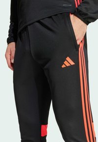 Pantalones deportivos negros con rayas verticales naranjas y logo de Adidas. Fabricados en una tela suave con bolsillos laterales con cremallera.
