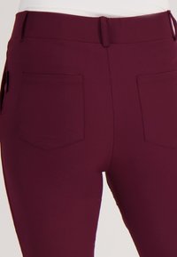 Bordeaux kleurige broek met een aansluitend ontwerp, hoge tailleband en twee achterzakken. Soepele textuur met subtiele stikseldetails.
