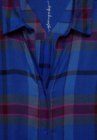 Blue plaid shirt featuring a button-down collar, a front placket with a red button, and alternating dark and light blue stripes.