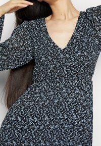 Robe noire avec motif floral bleu, décolleté en V, longues manches bouffantes et taille froncée. Tissu léger et texturé ajoutant de la dimension.