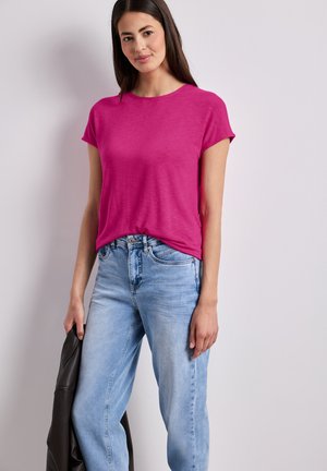 Nuori nainen, jolla kirkkaanpunainen lyhythihainen paita ja vaaleansiniset farkut, pitää mustaa takkia yksiväristä vaaleaa taustaa vasten.