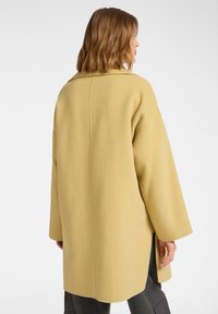 Cappotto di lana giallo con vestibilità rilassata, maniche ampie e apertura sulla schiena. Presenta un colletto a coscia e una superficie liscia.