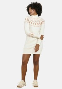 Abito in maglia crema con motivi geometrici in rosa chiaro e marrone. Presenta un colletto alto e maniche lunghe, terminando sopra il ginocchio.