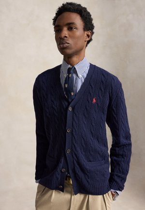 Homme portant un cardigan en maille torsadée bleu marine sur une chemise rayée et une cravate à motifs avec un pantalon beige sur un fond neutre.