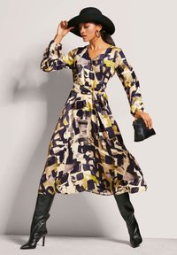 Langärmliges Kleid mit floralem Muster in Schwarz, Creme und Gelb, mit tailliertem Bund und fließendem Rock. Mit passendem schwarzen Hut.