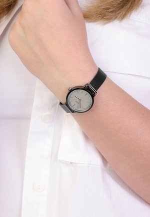 Schwarze Mesh-Uhr mit rundem silbernem Zifferblatt, minimalistischen Stundenmarkierungen und einer seitlichen Krone. Am Handgelenk über einem weißen Hemd getragen.