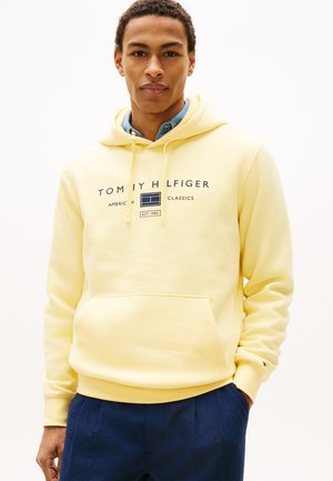 Jaunas vyras dėvi šviesiai geltoną Tommy Hilfiger džemperį su logotipo užrašu, po juo mėlyną marškinius ir tamsiai mėlynas kelnes, stovintis viena ranka kišenėje.