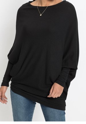 Femme portant un haut noir ample à manches longues côtelées avec un jean bleu et un petit collier pendentif rond sur un fond uni.