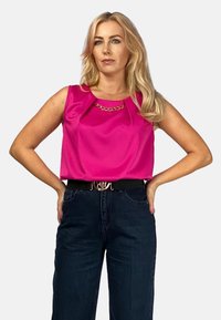 Bezrękawnik w jasnoróżowym kolorze z łańcuszkiem w dekolcie, w zestawie z ciemnoniebieskimi, wysokimi spodniami dżinsowymi i czarnym paskiem.