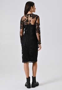 Robe en dentelle noire avec manches longues, motif floral, silhouette cintrée et détail à encolure en trou de serrure à l'arrière. Portée avec des bottines noires.