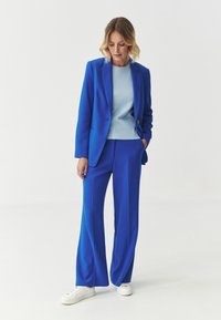 Blue tailored blazer and matching wide-leg trousers made from smooth fabric. Underneath, a light blue crew-neck top. White sneakers complete the look.