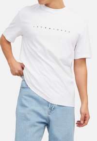 Witte katoenen T-shirt met ronde hals, korte mouwen, met zwarte tekst "JACK&JONES" op de borst, gecombineerd met lichtblauwe jeans.