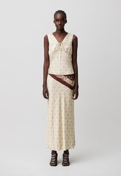 Donna che indossa un top beige senza maniche e una lunga gonna abbinata con dettaglio di sciarpa marrone a fantasia e tacchi neri con lacci, in piedi su sfondo bianco.
