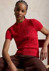 Kobieta z warkoczem, ubraną w czerwony, krótki sweter z wzorem warkoczy, ciemne spodnie, siedząca z jedną ręką opartą na krześle.