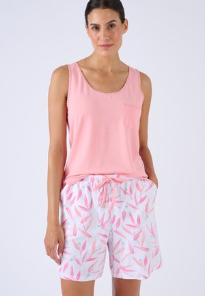 Femme portant un top rose sans manches avec poche avant et un short blanc avec motifs de feuilles roses, mains dans les poches, sur fond uni.