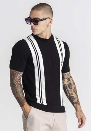 Man met kort haar en tatoeages op beide armen, die een zonnebril draagt, een zwart kort mouw shirt met witte verticale strepen en een lichte broek.