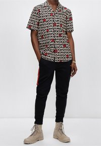 Maglietta a maniche corte con stampa nera, beige e rossa con il testo "HUGO"; abbinata a pantaloni joggers neri e stivali beige.