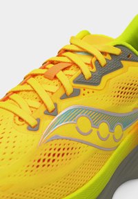 Φωτεινό κίτρινο αθλητικό παπούτσι με κορδόνια πορτοκαλοκίτρινου χρώματος, γκρι λεπτομέρειες και μπλε-ασημένιο λογότυπο σε μια διάτρητη, αναπνέουσα επιφάνεια.