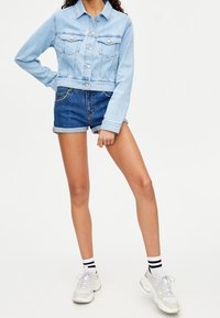 Persona vestida con una chaqueta de mezclilla azul clara, pantalones cortos de mezclilla azul oscura arremangados, zapatillas blancas y calcetines blancos con rayas negras, de pie contra un fondo blanco.