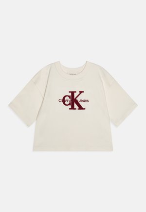 Camiseta corta de color crema con mangas cortas, presenta un logo bordado en color burdeos de "Calvin Klein Jeans" en la parte delantera. Textura de tela suave.