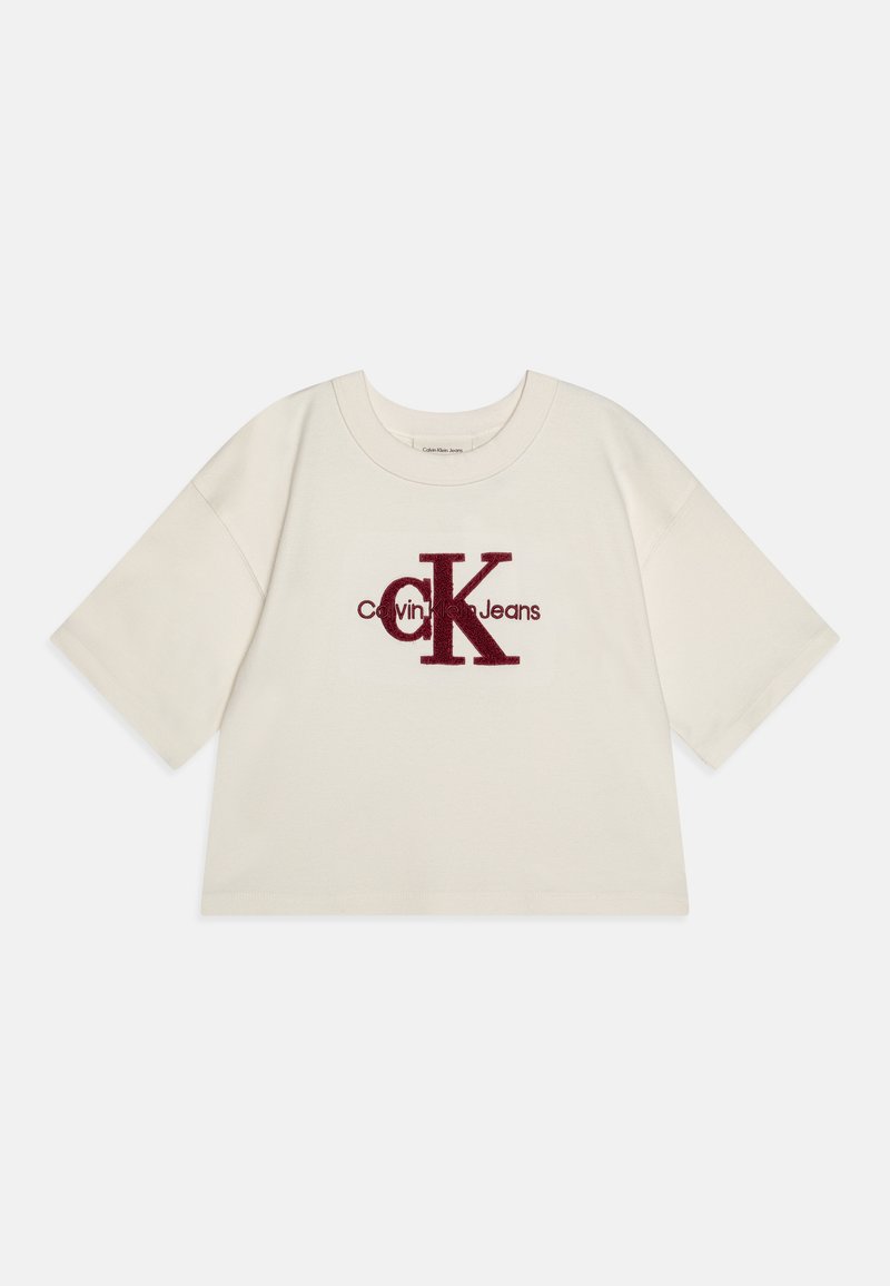 Camiseta corta de color crema con mangas cortas, presenta un logo bordado en color burdeos de "Calvin Klein Jeans" en la parte delantera. Textura de tela suave.
