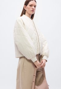 Λευκό μπουφάν bomber με καπιτονέ ύφασμα, φουσκωτά μανίκια και ριμπ μανσέτες, σε συνδυασμό με μπεζ φούστα και κρατώντας μια τσάντα χρώματος ανοιχτό ροζ.