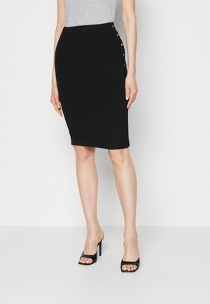 Femme portant une jupe crayon noire longueur genou avec des boutons latéraux, haut gris et des sandales noires à talons hauts, debout sur un sol blanc.