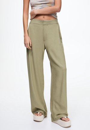 Woman wearing loose light olive pants, a cropped tank top, and white platform sandals, standing against white background.