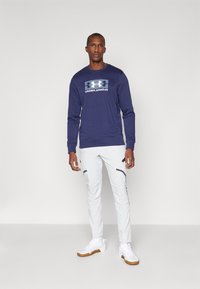 Marineblaues Langarm-Sweatshirt mit einem zentrierten weißen Logo, kombiniert mit weißen tapered Hosen mit schwarzen Reißverschluss-Taschen und Bündchen. Weiße Sneaker.