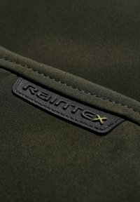 Tissu vert foncé avec un gros plan sur une étiquette noire cousue embossée du mot « Raimtex » et un petit « x » jaune sur le côté droit.