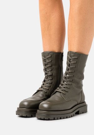 Buty zawiązywane na sznurowadła z oliwkowej skóry, o designie sięgającym do połowy łydki, z teksturowaną cholewką, solidną gumową podeszwą oraz suwakiem z boku dla łatwego zakładania.