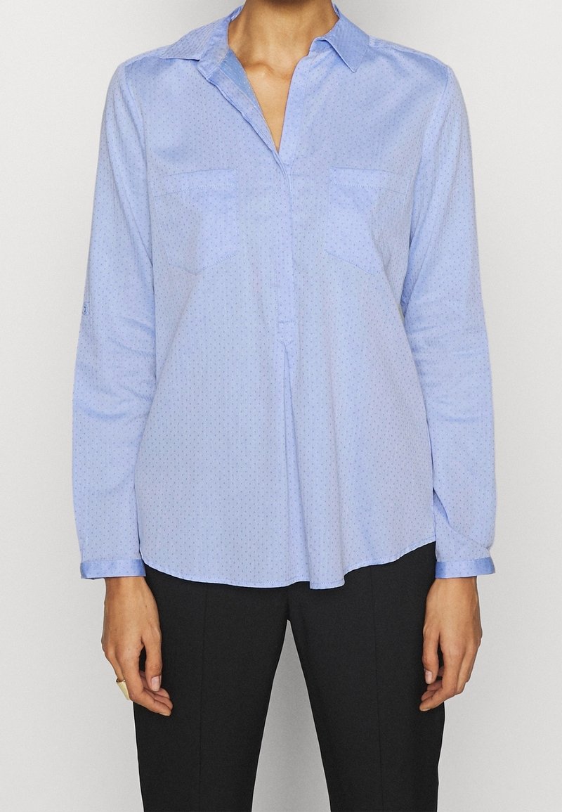 Femme portant un chemisier bleu clair à manches longues avec de petits pois et un pantalon noir, debout devant un fond uni.