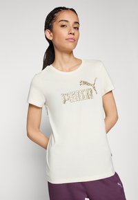 Camiseta de algodón color crema con el logo de "PUMA" en un patrón de animal dorado y un pequeño logo de un gato saltando, con mangas cortas y un cuello redondo.