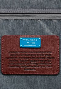 Piquadro Gepäckanhänger: dunkelgrauer, strukturierter Stoff, kastanienbraunes Lederetikett mit geprägt Text, blaue Metall-Logo-Plakette, rote Nähakzente.