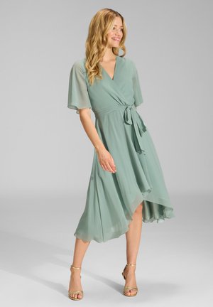 Femme portant une robe portefeuille vert clair avec des manches volantes et un ourlet asymétrique, associée à des talons dorés à brides, debout sur un fond uni.