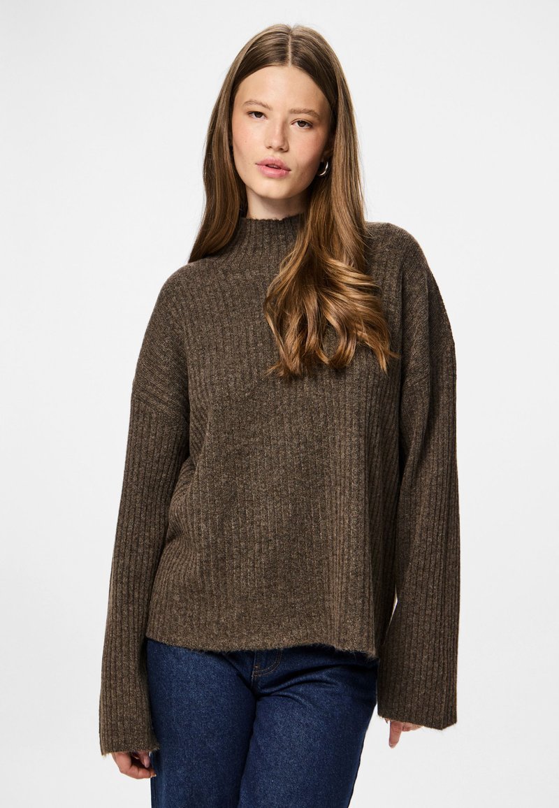 Femme jeune avec de longs cheveux bruns portant un pull marron côtelé et un jean bleu, se tenant devant un fond blanc uni.